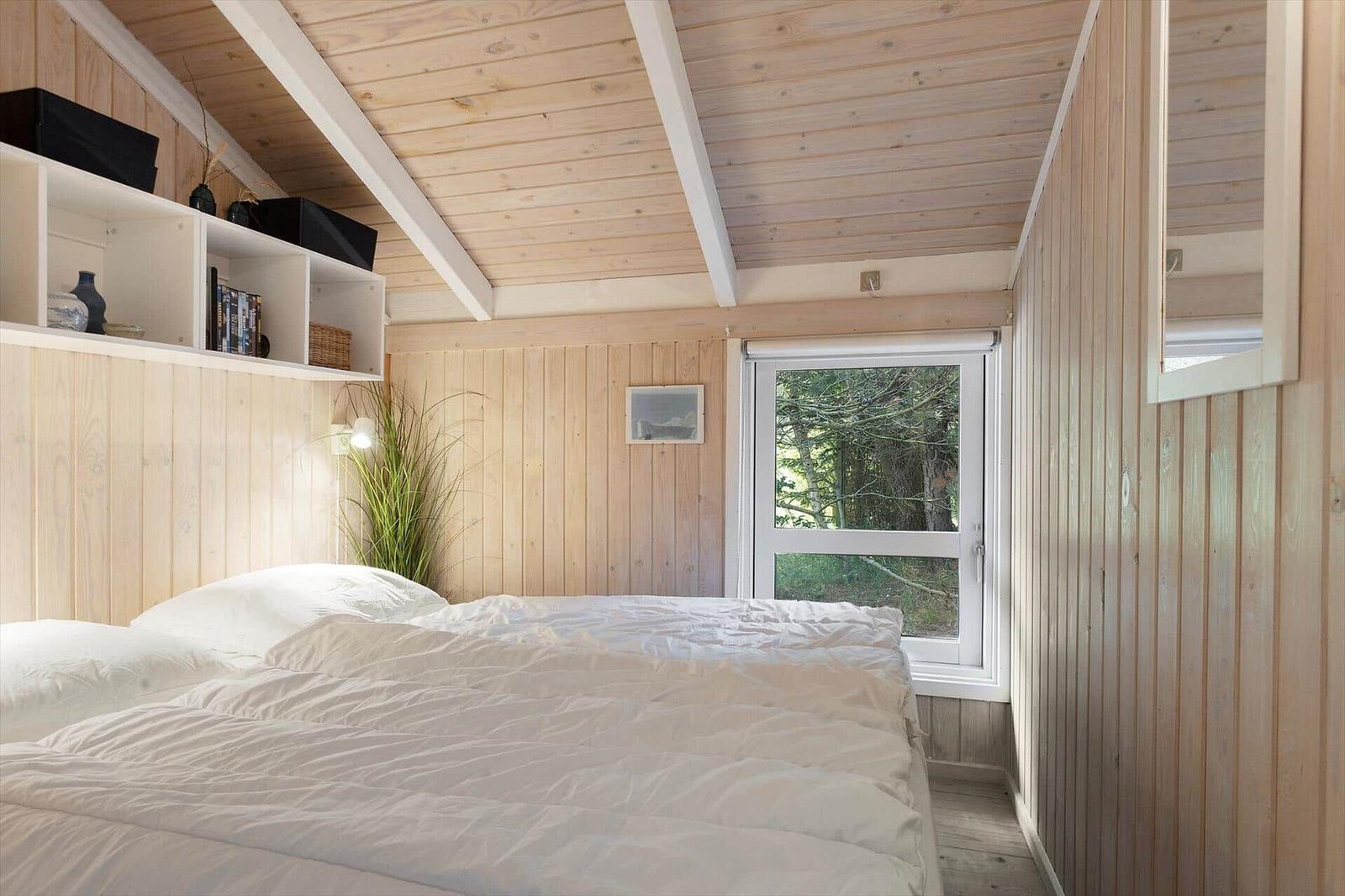 Bedroom with wooden walls and ceiling beams, bed with white bedding, window with view of forest.