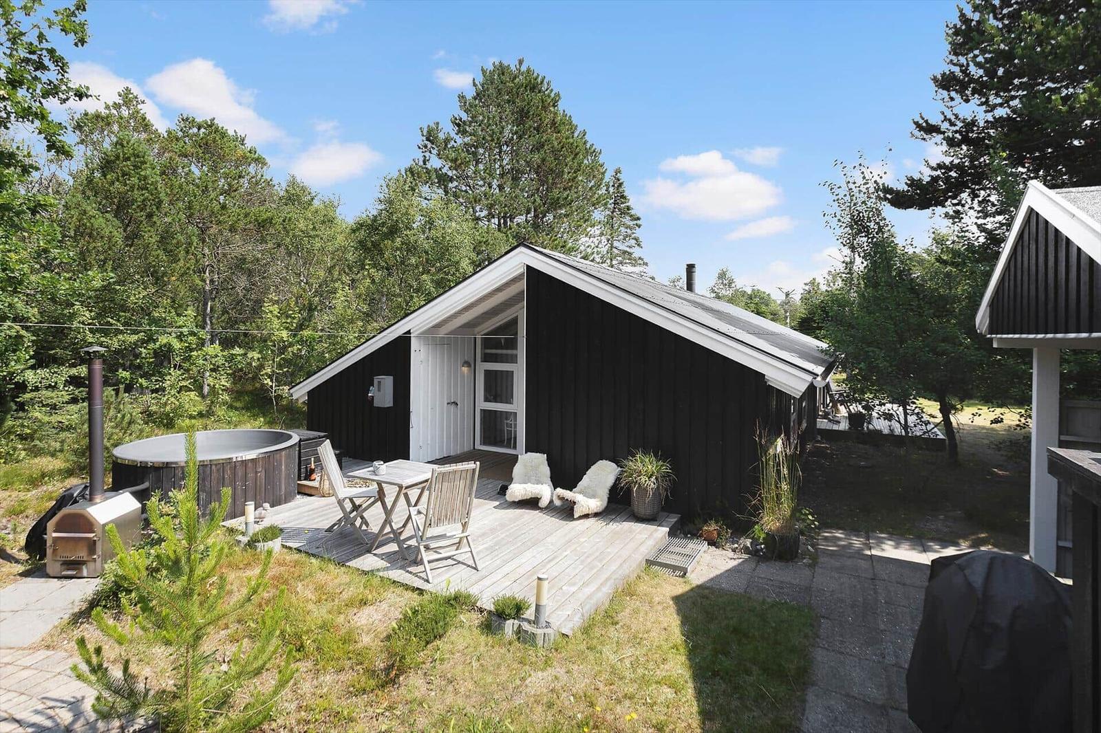 Black wooden house with terrace, hot tub, and grill area in the woods.