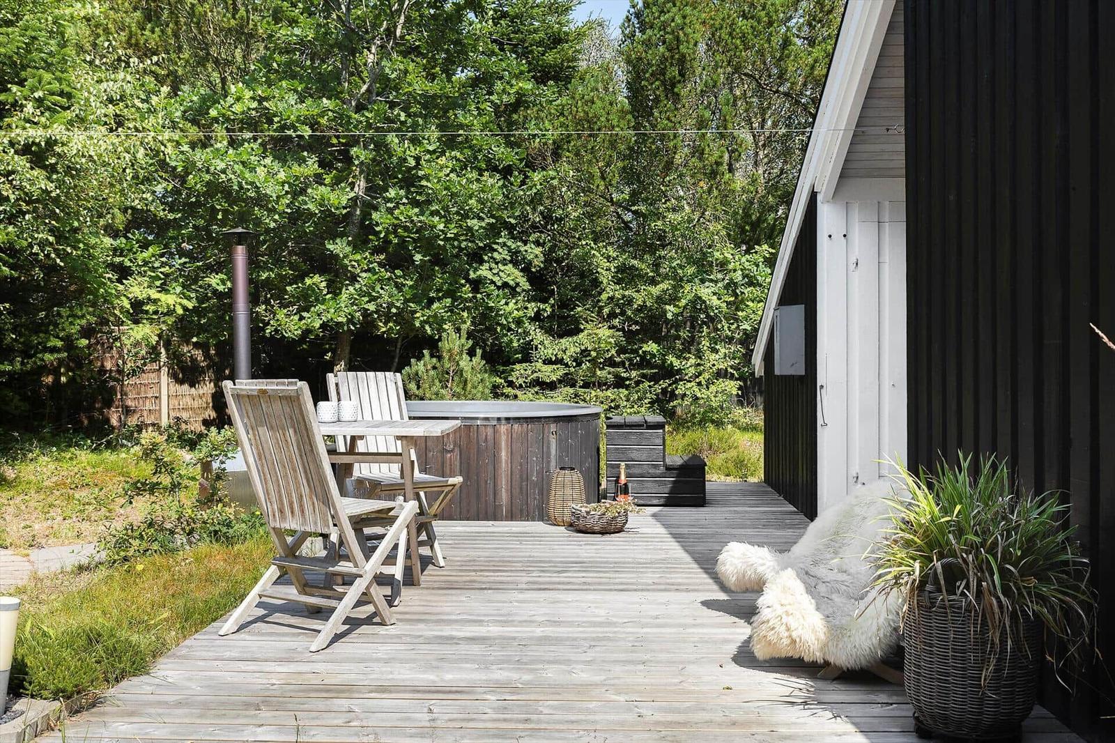 Deck with wooden table, chairs, and hot tub. Black wall and green trees in background.