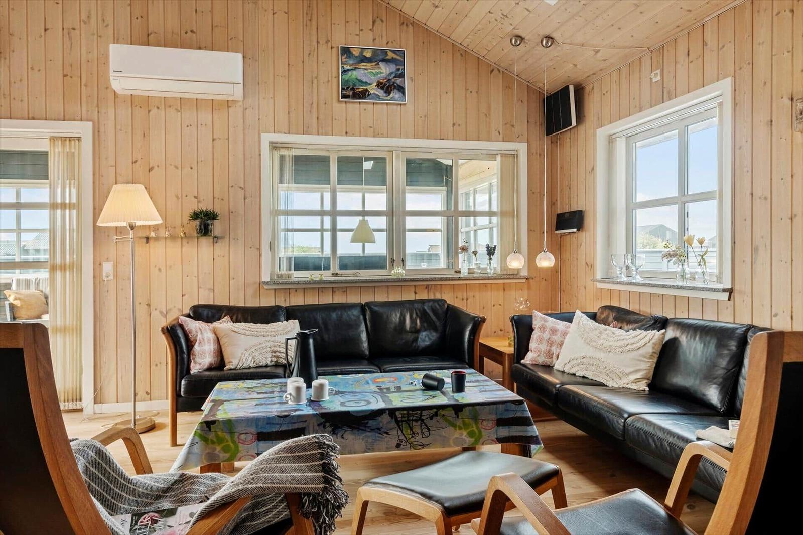 Wohnzimmer mit zwei schwarzen Sofas, Holzwand und Fenstern. Tisch mit Tischdecke und Stühle.