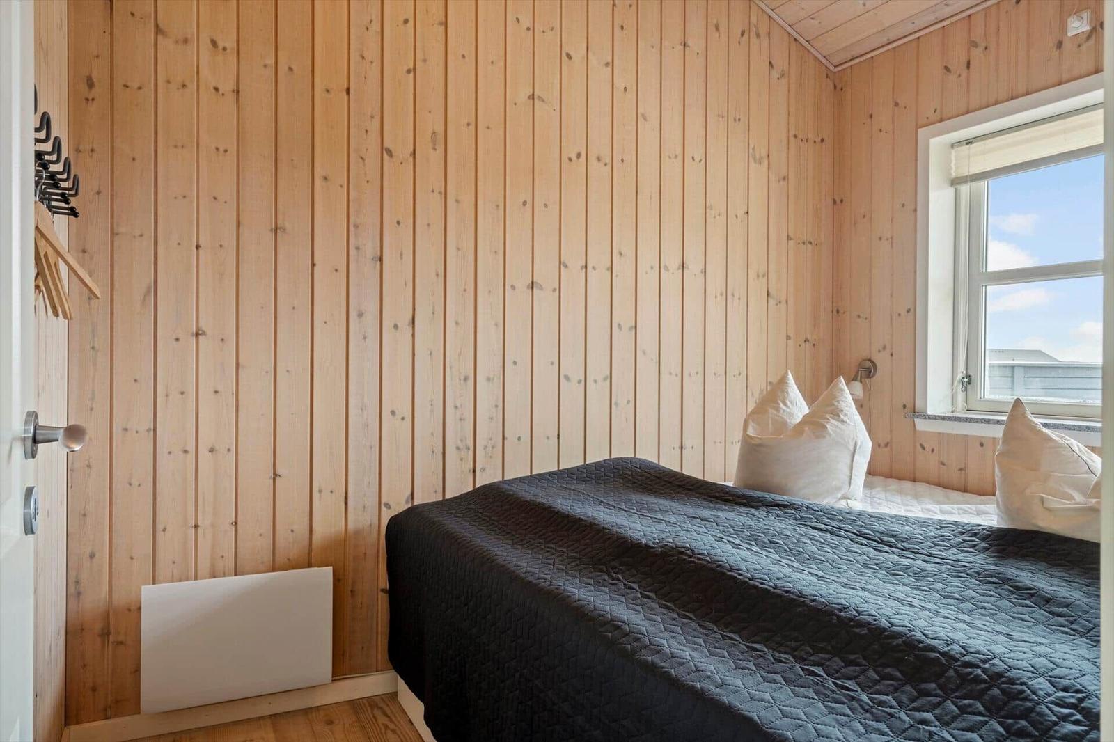 Schlafzimmer mit Holzwand, Bett mit dunkler Bettdecke und Fenster.