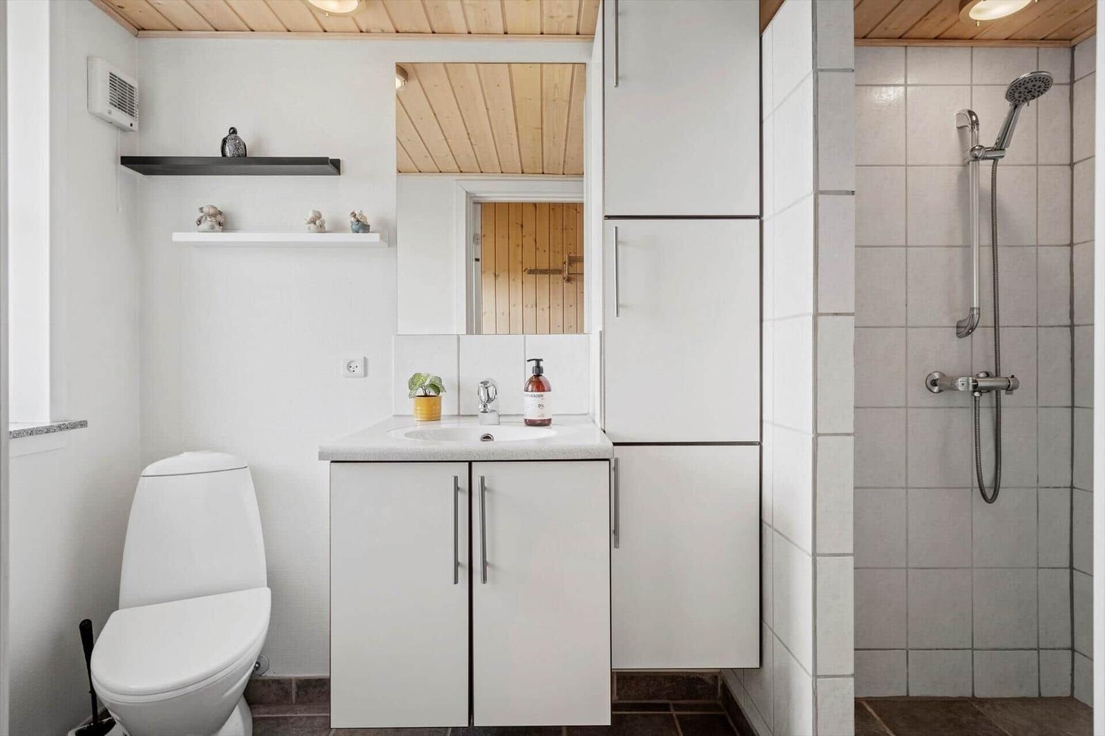 Badezimmer mit Dusche, Waschbecken und Toilette. Holzdecke und weiße Schränke.