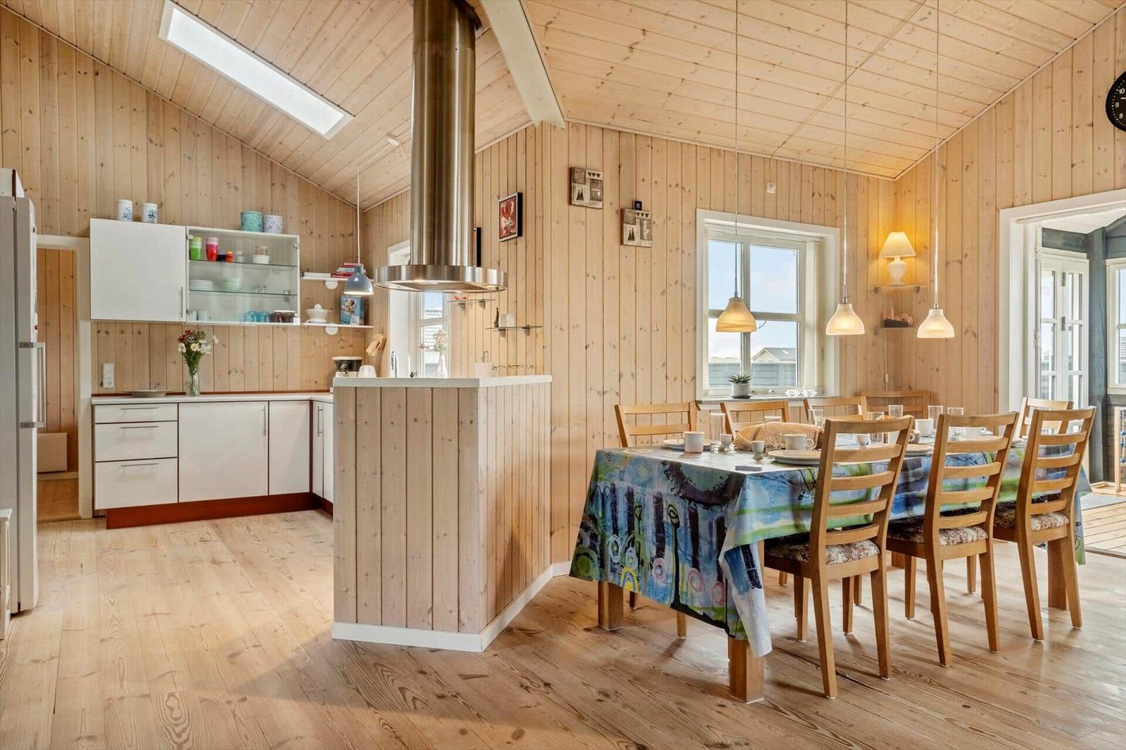 Küche und Essbereich mit Holzwand und -boden, Tisch mit Tischdecke und Stühlen, Fenster und Deckenleuchten.