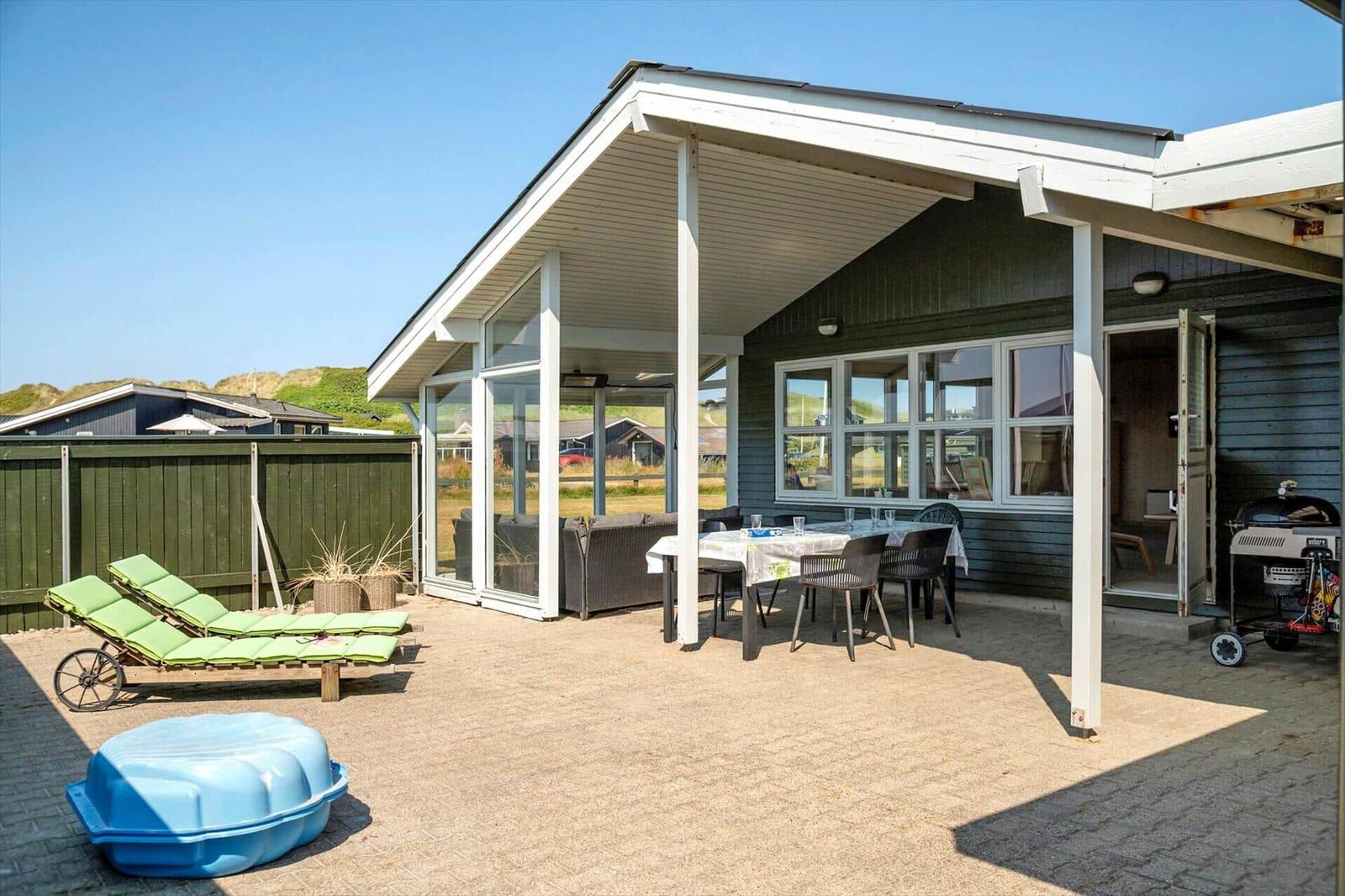 Terrasse mit Tisch, Stühlen, Liegestühlen und Grill. Haus mit Glasfront und Holzwand.