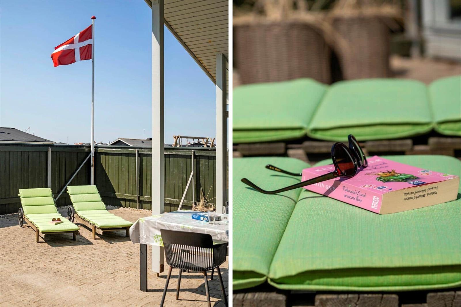 Terrasse mit Liegen, Tisch und dänischer Flagge. Grün gepolsterte Sitzmöbel und ein Buch.