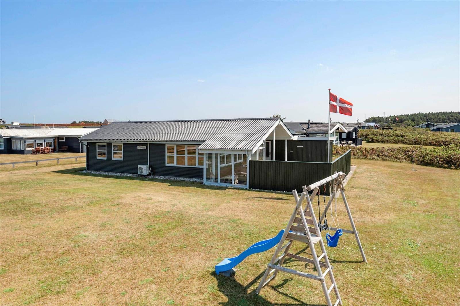 Ein dunkelgrauer Ferienhaus mit Spielplatz und dänischer Flagge auf grünem Grund.