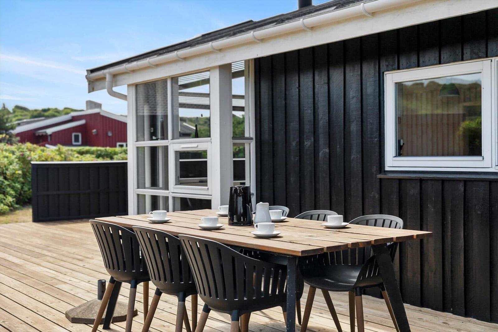 Wooden table with six chairs on a terrace in front of a house with black siding.