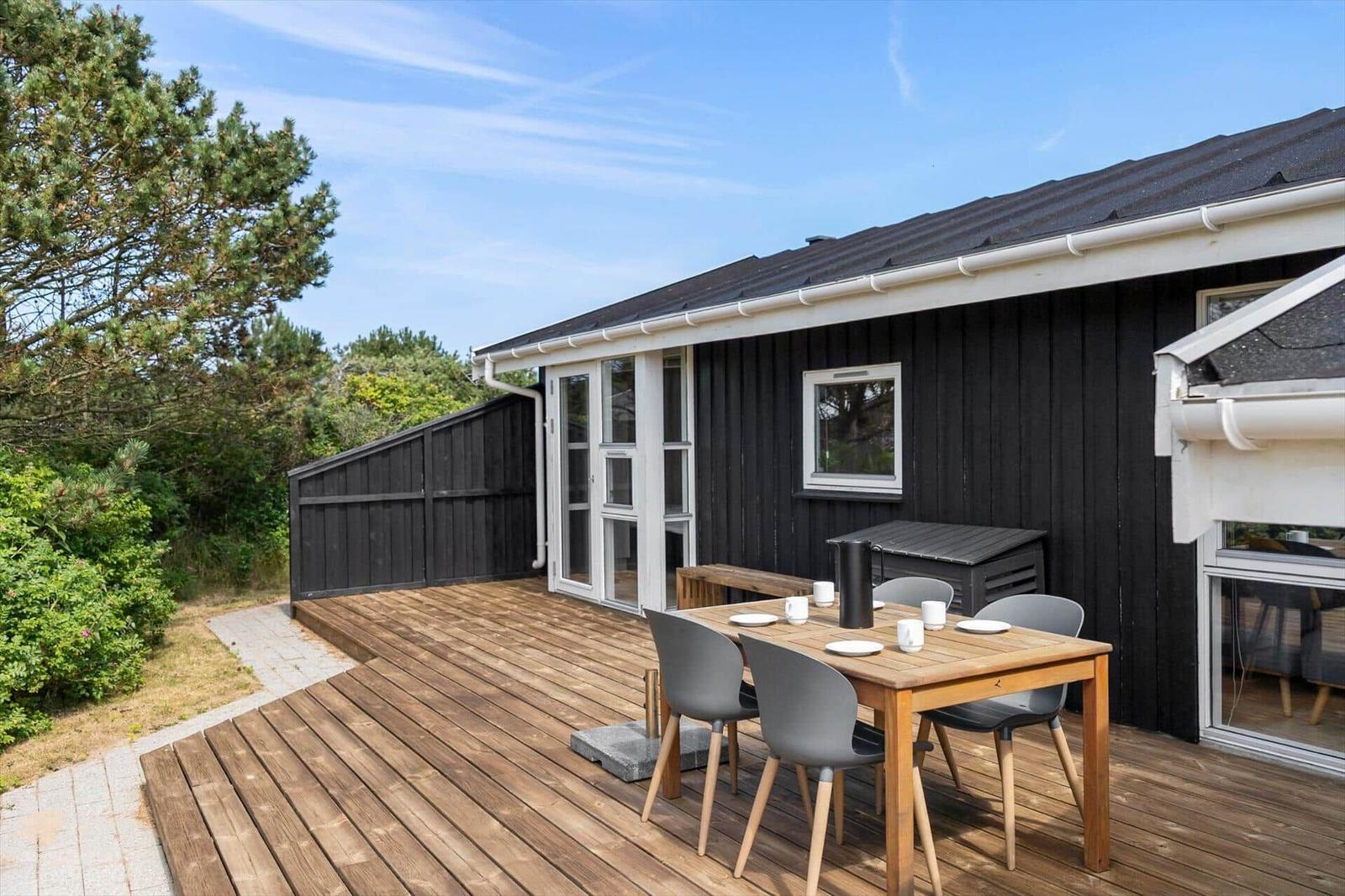 Deck with wooden table, four chairs, and grill. Black wooden wall and windows. Greenery behind the house.
