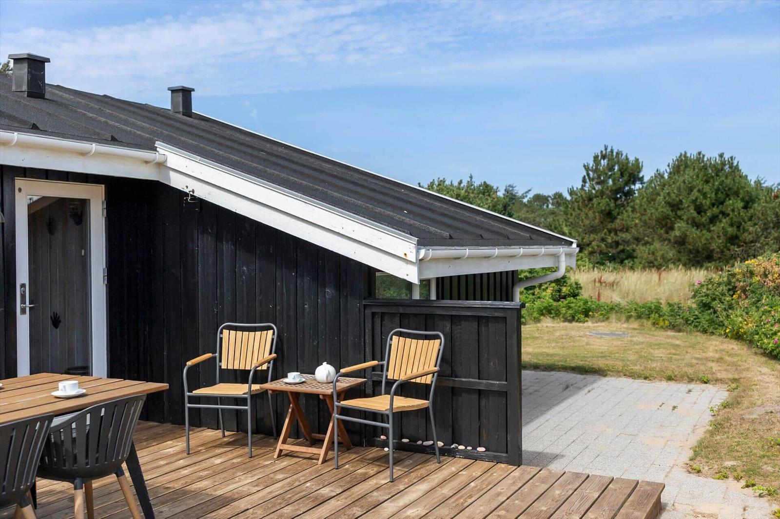 Wooden terrace with table and chairs in front of a black-painted house with gable roof.
