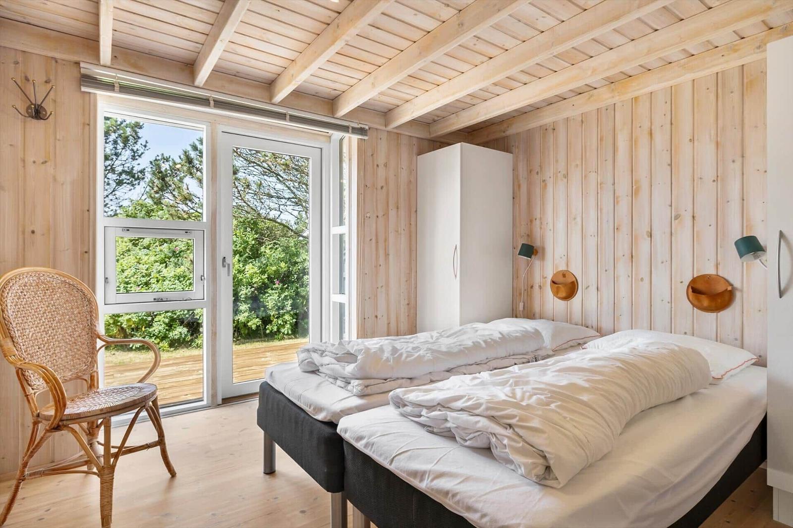 Two beds, wooden walls, window with view of green, chair and wardrobe.