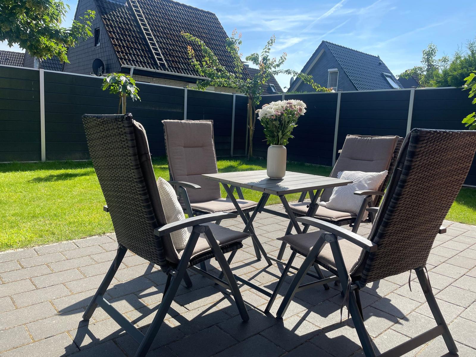 Terrasse mit Tisch und vier Stühlen, Blumen in Vase, grüner Rasen und Zaun im Hintergrund.
