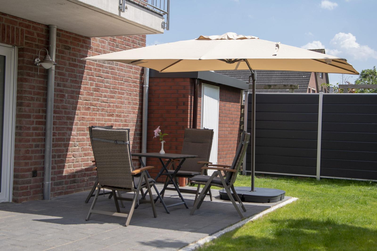Terrasse mit Tisch, Stühlen und Sonnenschirm. Hintergrund: Backsteinmauer und schwarze Holzumzäunung.