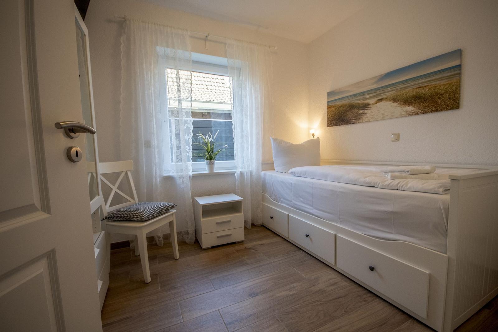 Ein Schlafzimmer mit weißem Bett, Nachttisch und Stuhl. An der Wand hängt ein Bild.