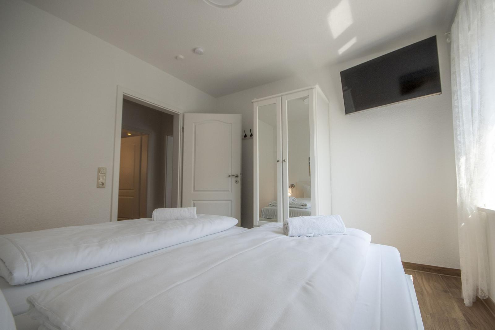 Schlafzimmer mit weißem Bett, Spiegelschrank und Wandfernseher.