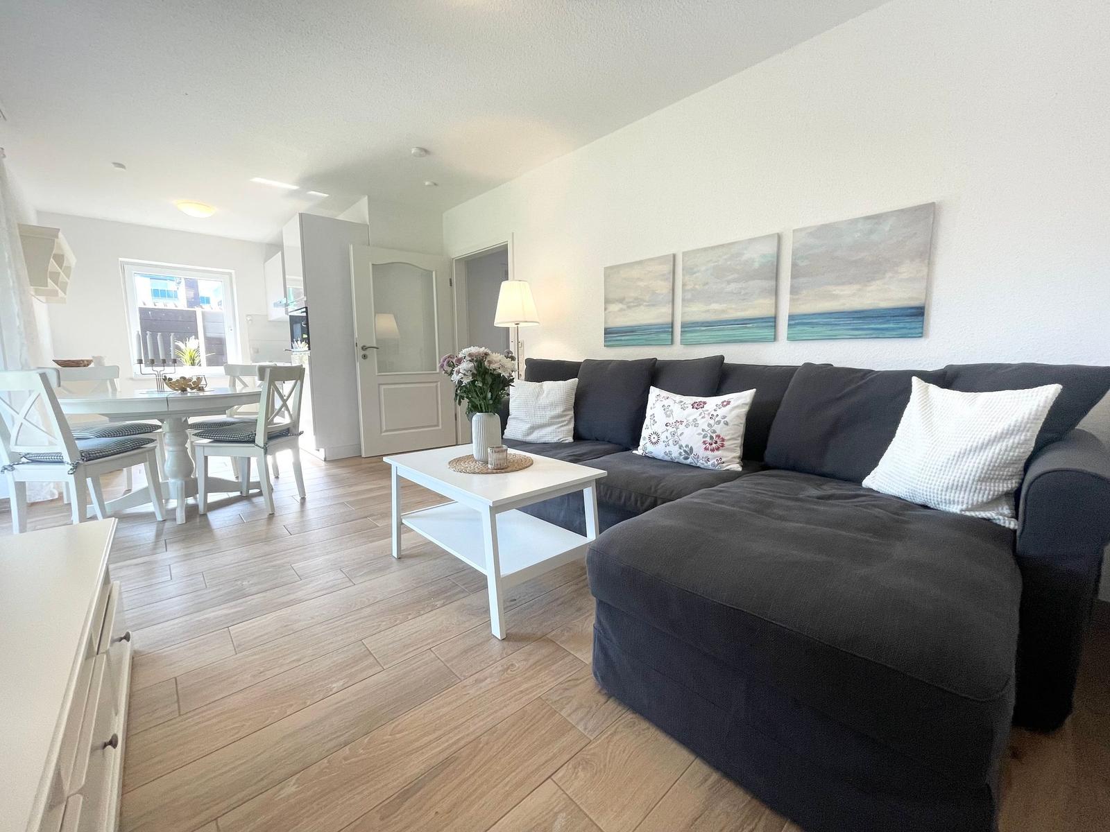 Ein Wohn- und Essbereich mit grauem Sofa, weißem Tisch und Wandbildern.