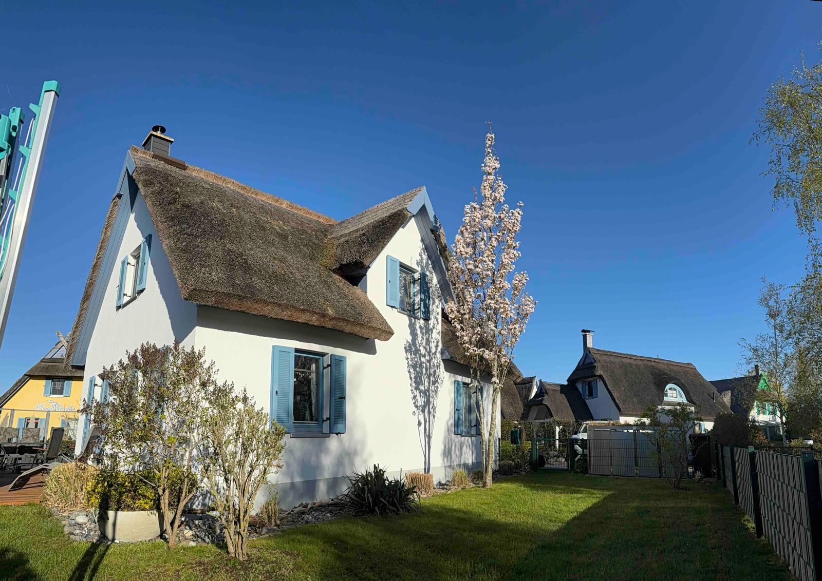Wohnhaus mit Strohdach und blauen Fensterläden, umgeben von Grünfläche und Blumen.