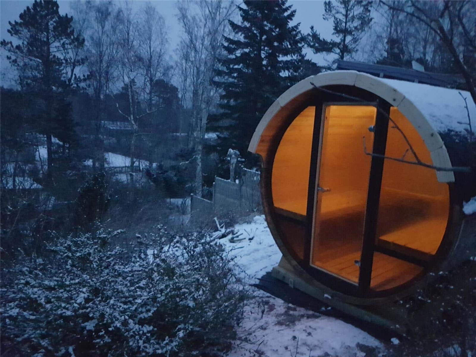 Holzsauna im Freien mit warmem Licht, umgeben von Schnee und Bäumen.