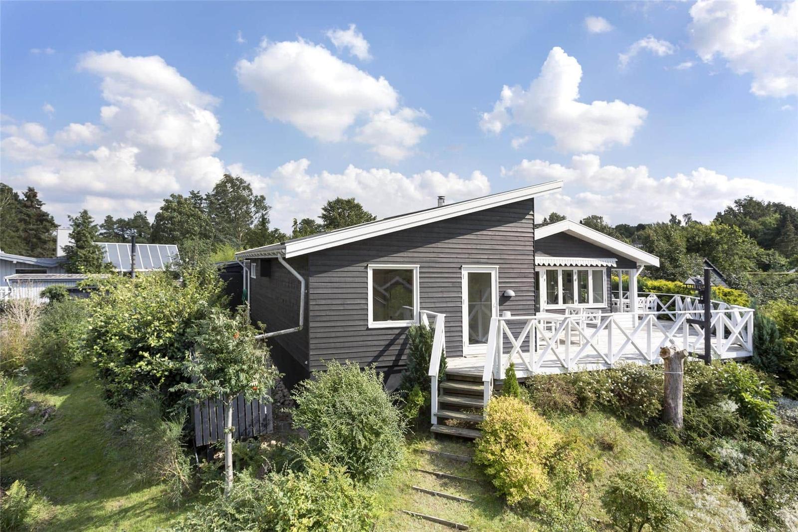 Ein graues Holzhaus mit weißer Veranda und Garten. Hintergrund: Bäume und blauer Himmel.