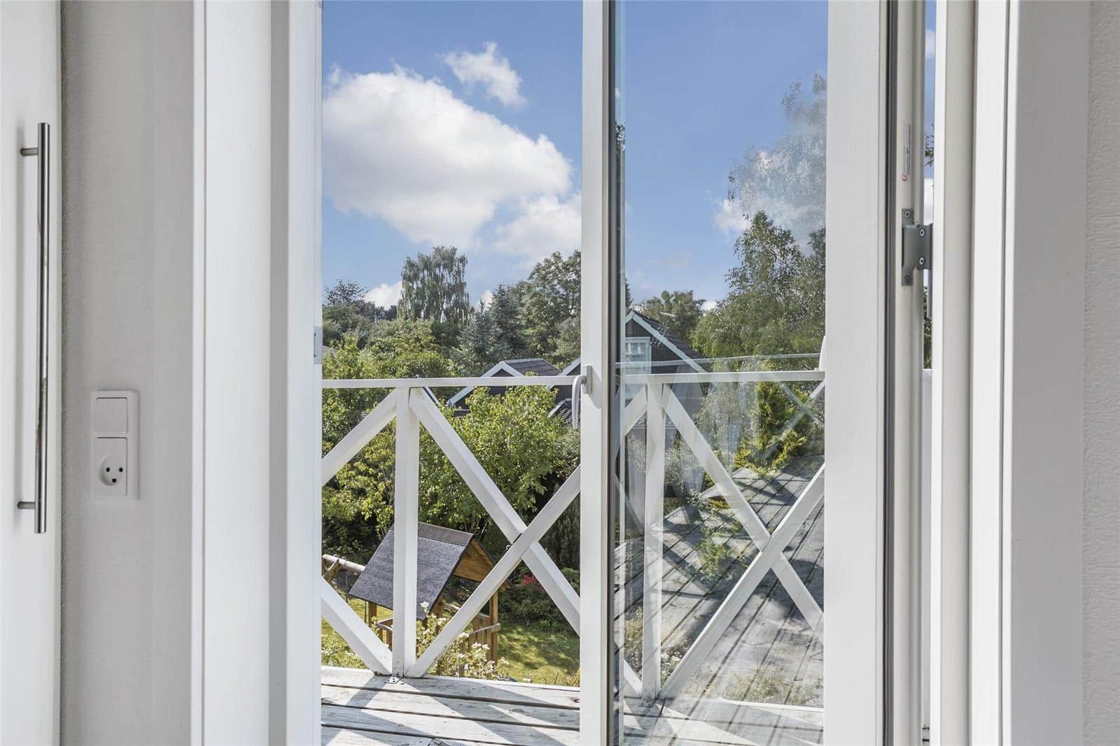 Durch die Glastüren blickt man auf eine Terrasse mit weißem Geländer und grüne Bäume im Hintergrund.