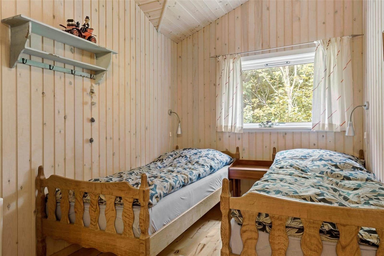 Two single beds with wooden frames, wall shelf, and window with view of forest.
