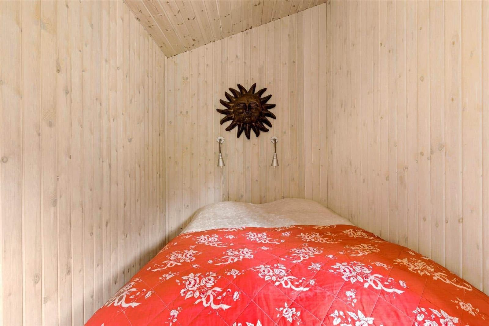 Bedroom with wood paneling, bed with red floral patterned bedspread, and sun artwork on the wall.
