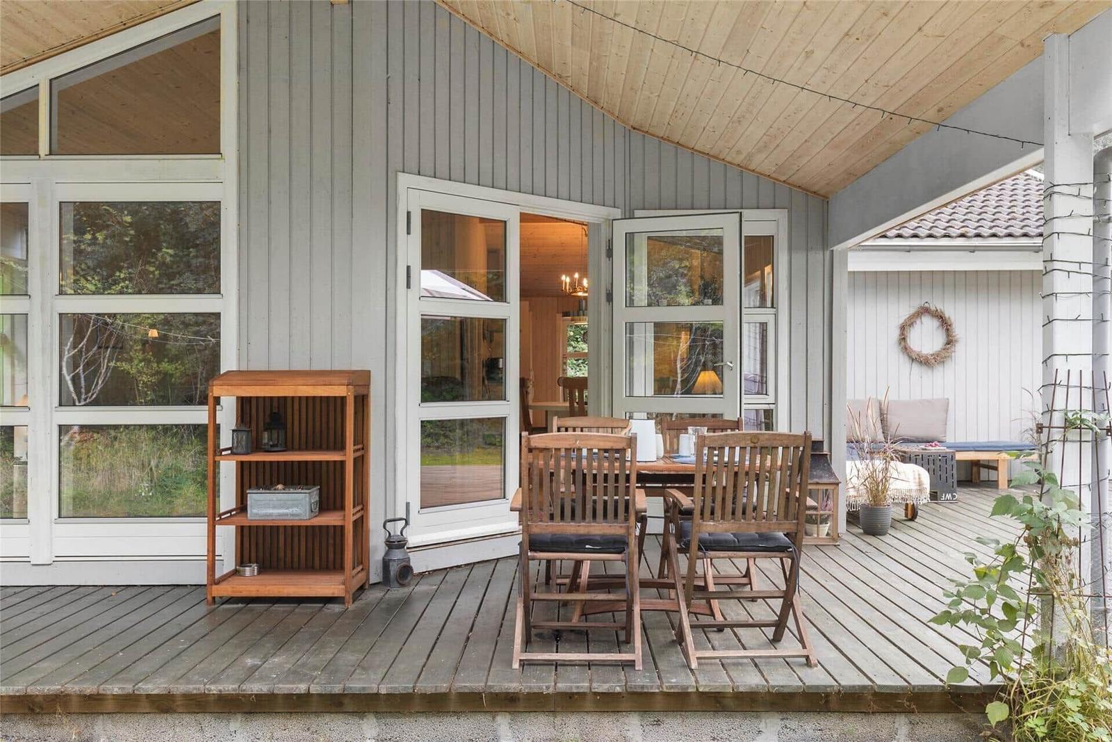 Deck with wooden table, chairs, and shelf. Entrance to apartment. Background: garden area.