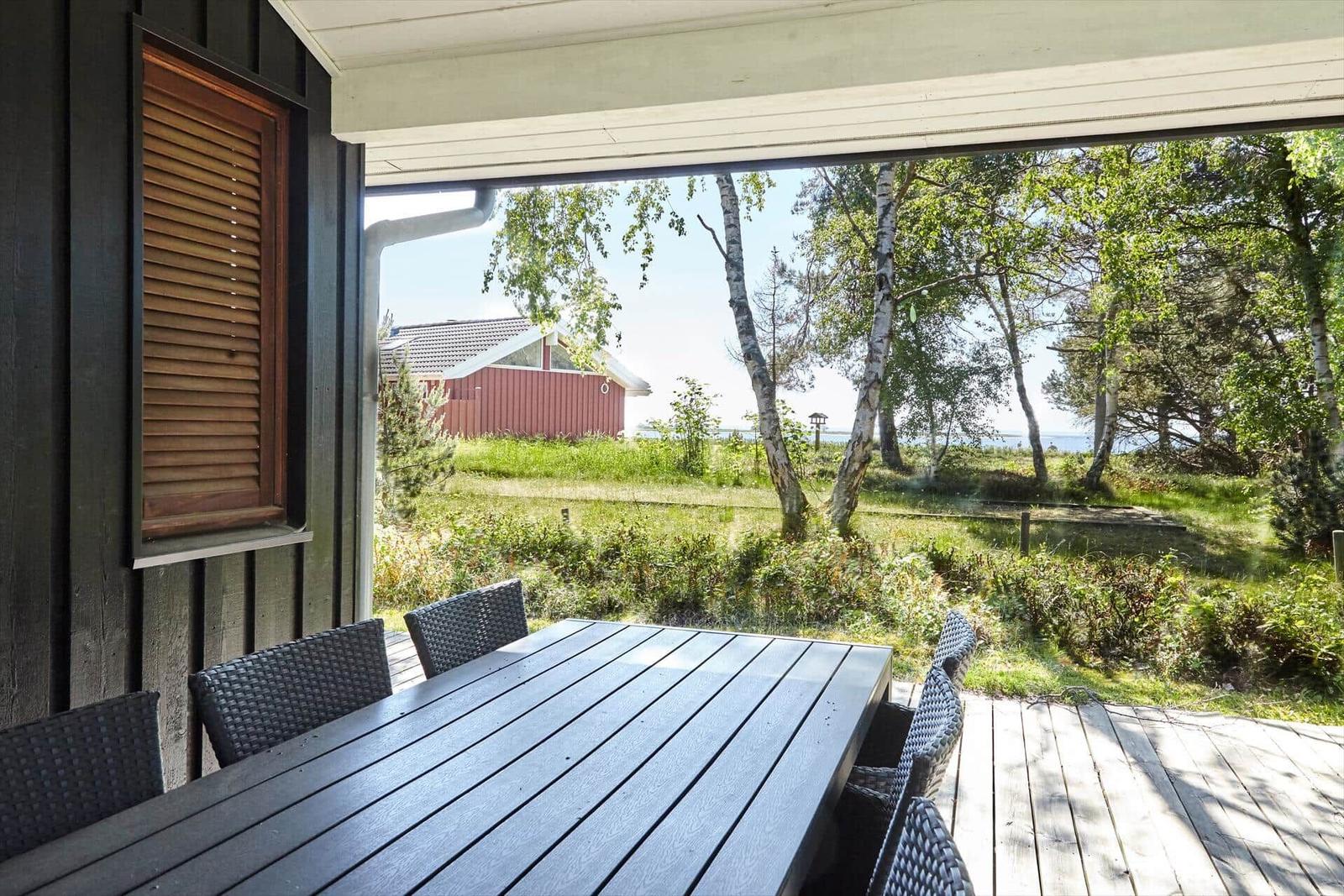Terrasse mit Grill, Liegestühlen und Tisch. Holzwand und Blick auf Bäume.
