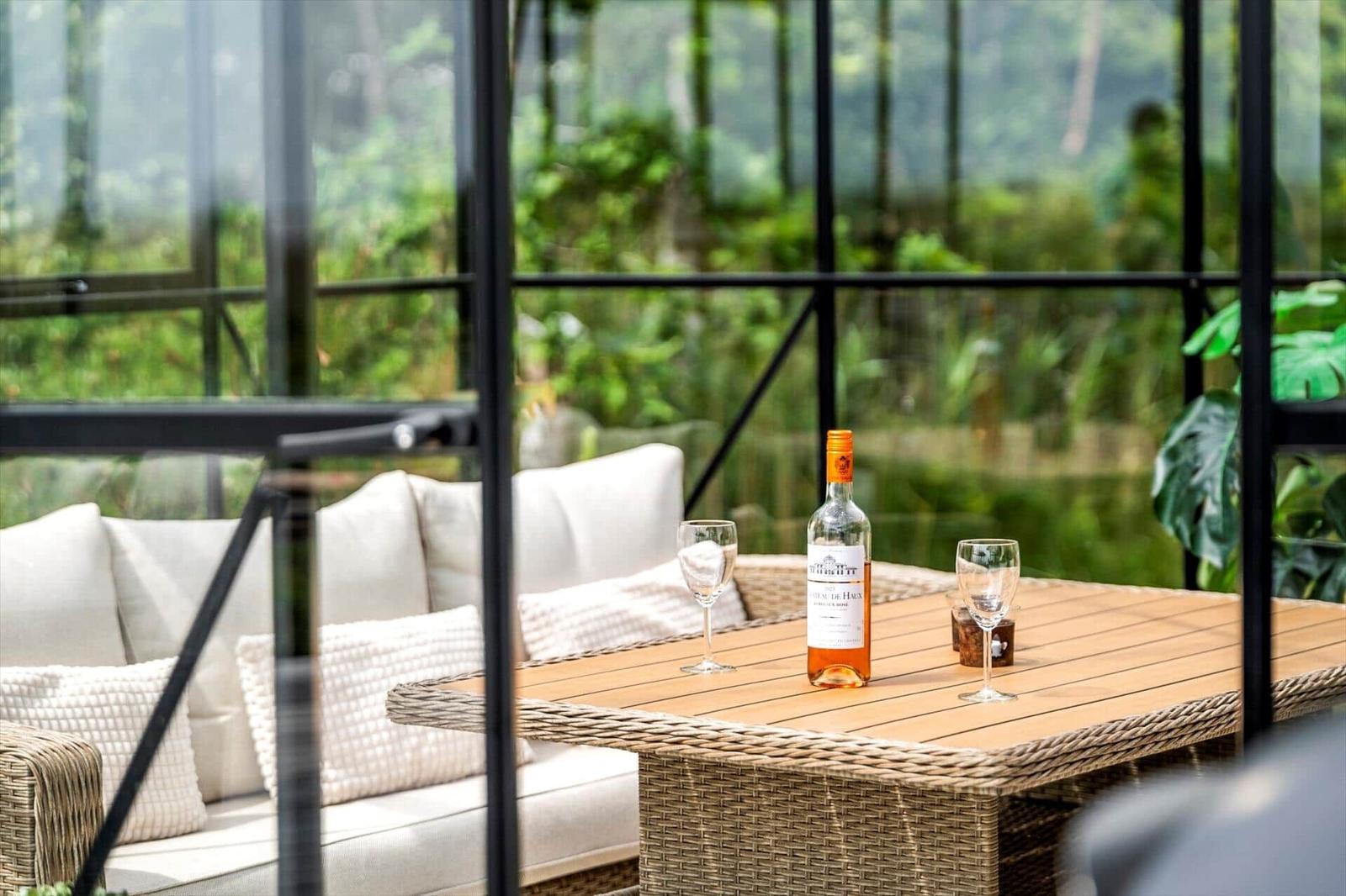 Terrasse mit Glasgarten, Rattanmöbeln und rotem Liegestuhl. Hintergrund: Grasland und Bäume.
