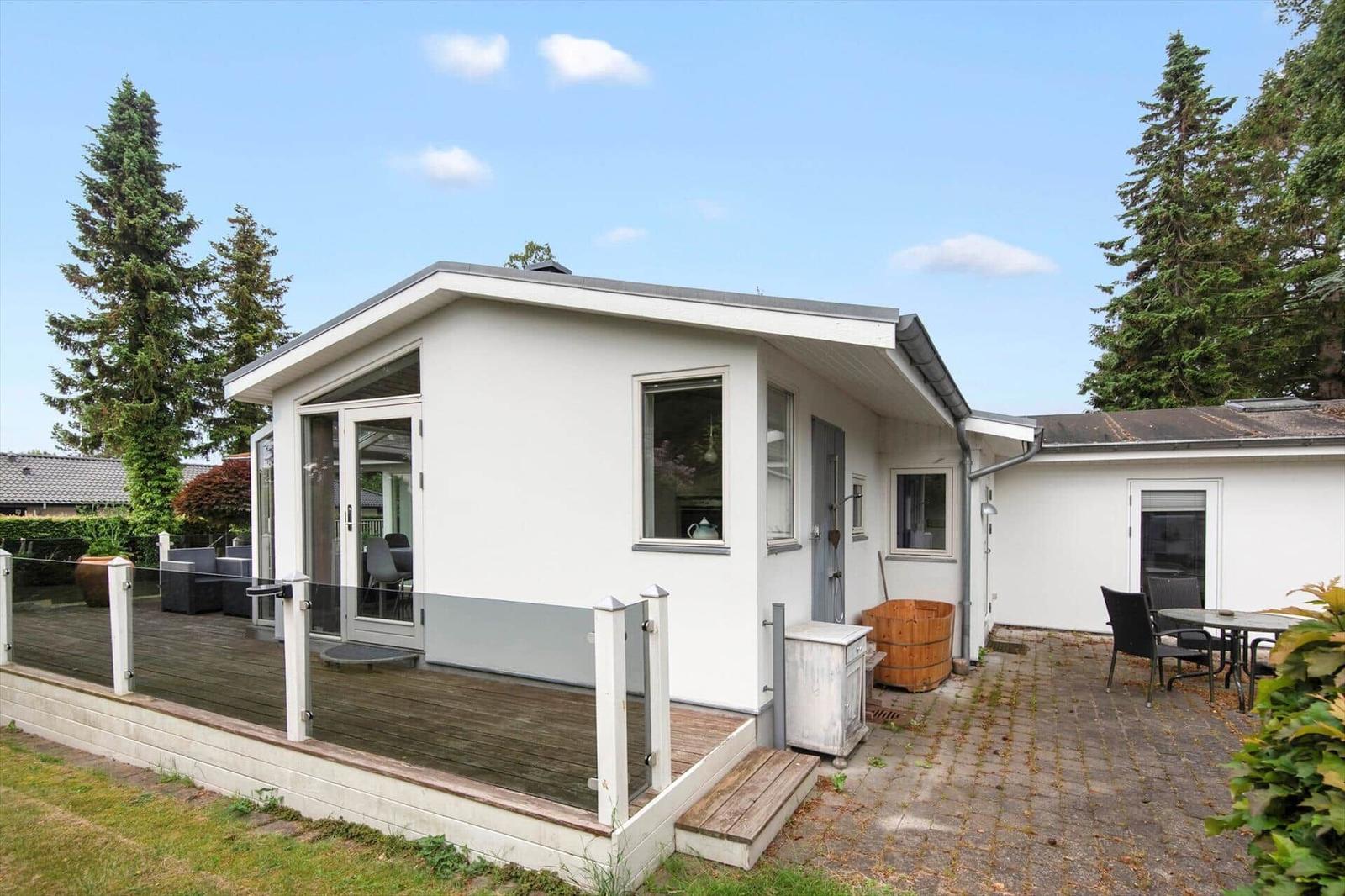 Moderne Villa mit überdachtem Balkon und großem Garten. Holzterrasse und Bäume umgeben das Haus.