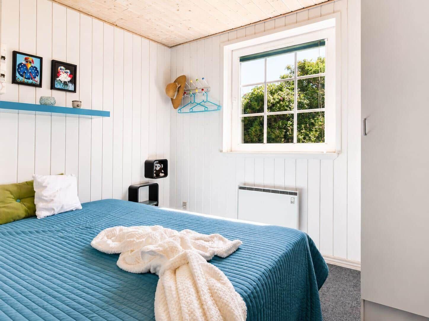 Schlafzimmer mit blauem Bett, Fenster und weißen Wänden.