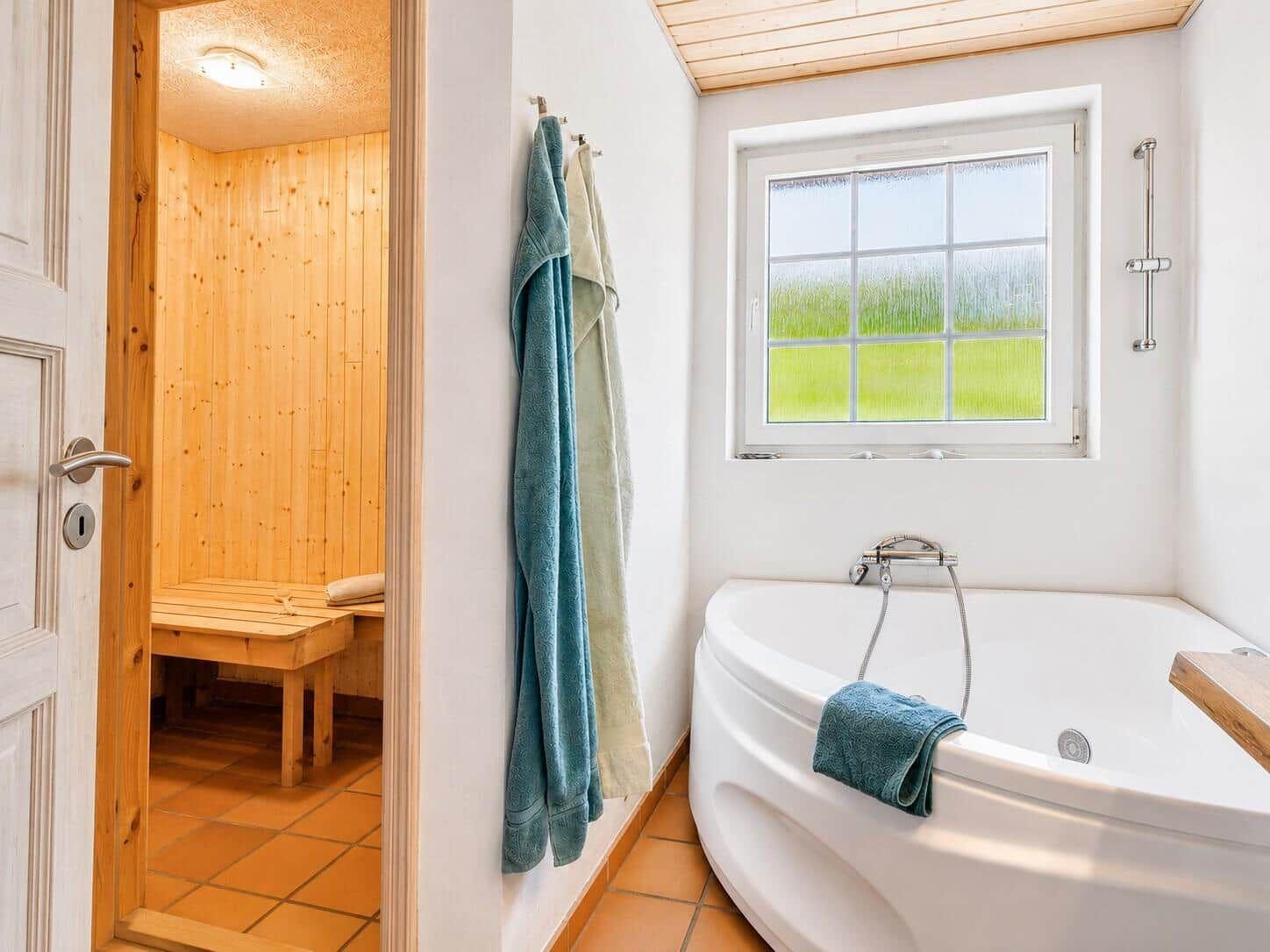 Badezimmer mit Badewanne und Sauna. Fenster mit grünem Blick. Handtücher an der Wand.