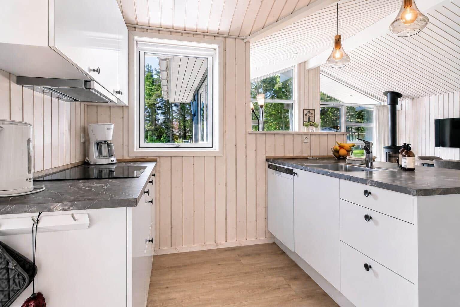 Küche mit weißen Schränken, dunklem Arbeitsplatz und Holzdielen. Fenster zeigen grüne Bäume.