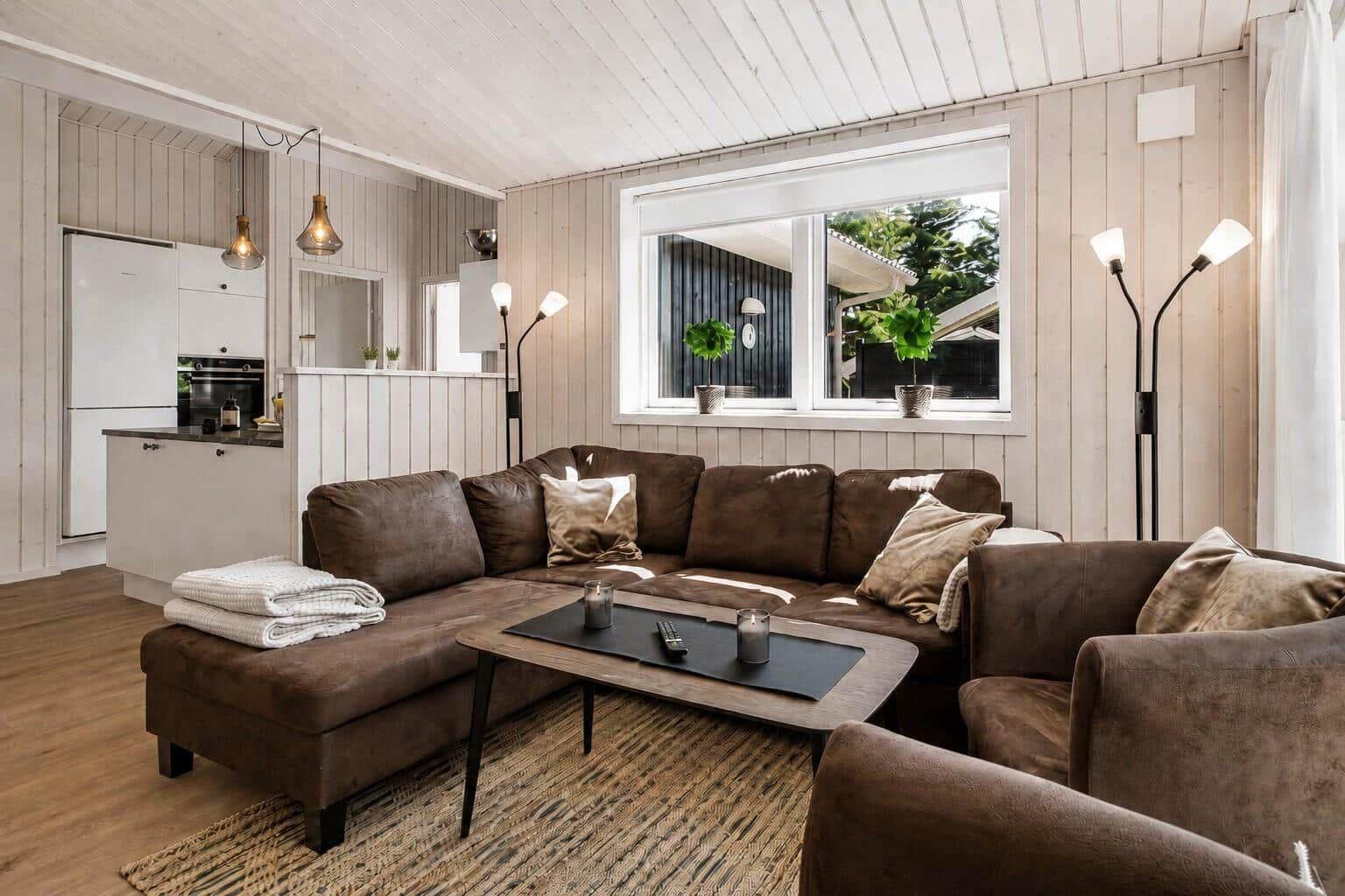 Wohnzimmer mit braunem Sofa, Tisch und Fenster. Küche im Hintergrund.