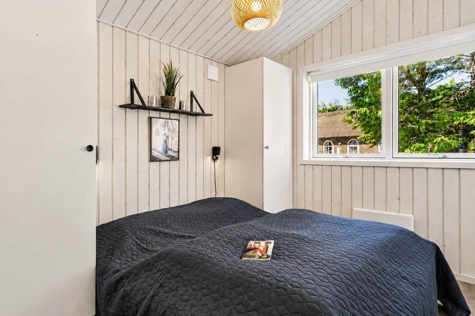 Schlafzimmer mit Bett, Wandregal und Fensterblick auf grüne Bäume.