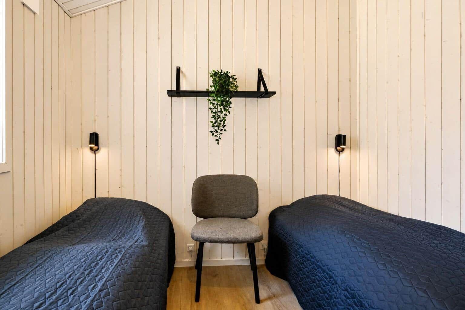 Zwei Betten mit dunklen Bezügen, Stuhl und Pflanze an der Wand.