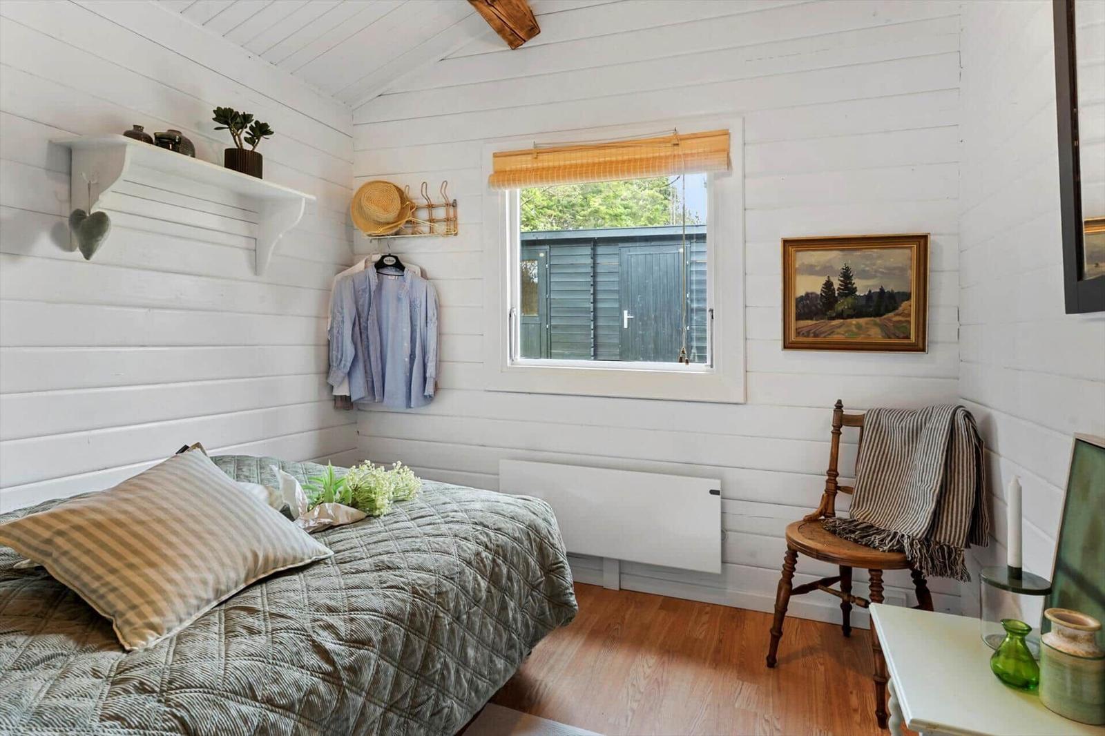 Schlafzimmer mit Bett, Wandleuchte und Fenster. Holzwände und ein Stuhl sind sichtbar.
