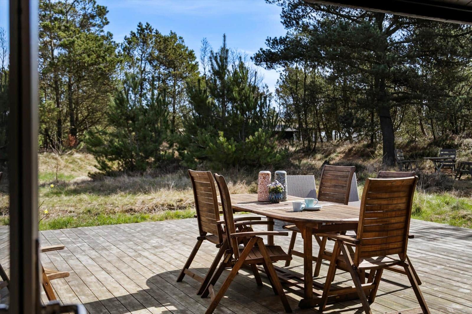 Ein dunkles Holzhaus mit Grasdach steht unter hohen Bäumen. Vor dem Haus befindet sich eine Terrasse.
