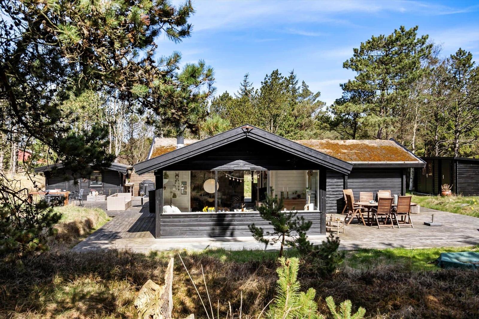 Holzterrasse mit Tisch und Stühlen vor einem Haus mit Moosdach und großem Fenster.
