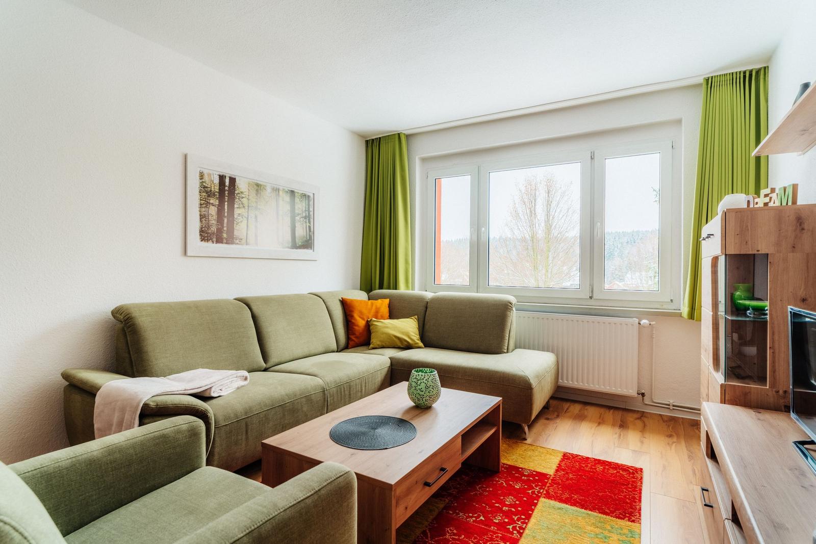 Ein Wohnzimmer mit grünem Sofa, Holztisch und Fensterblick auf Wald.