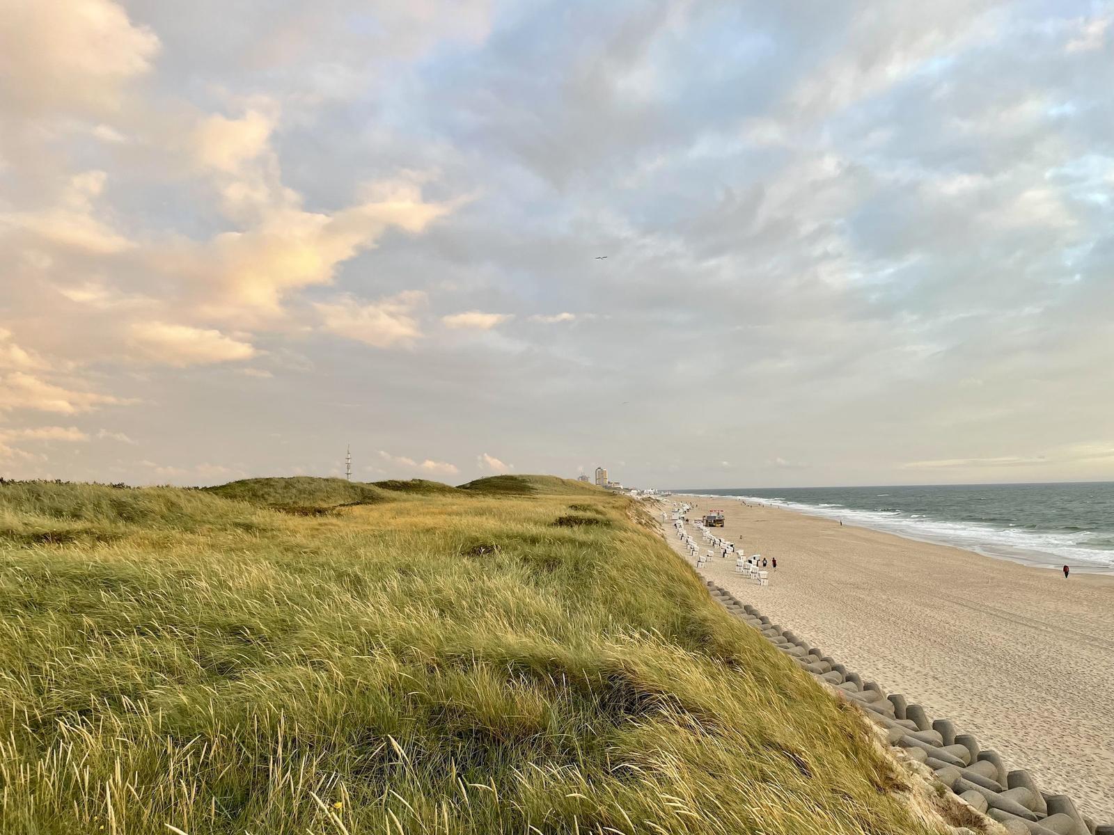 Strand mit Sand, Strandkörben und Holzsteg unter blauem Himmel.