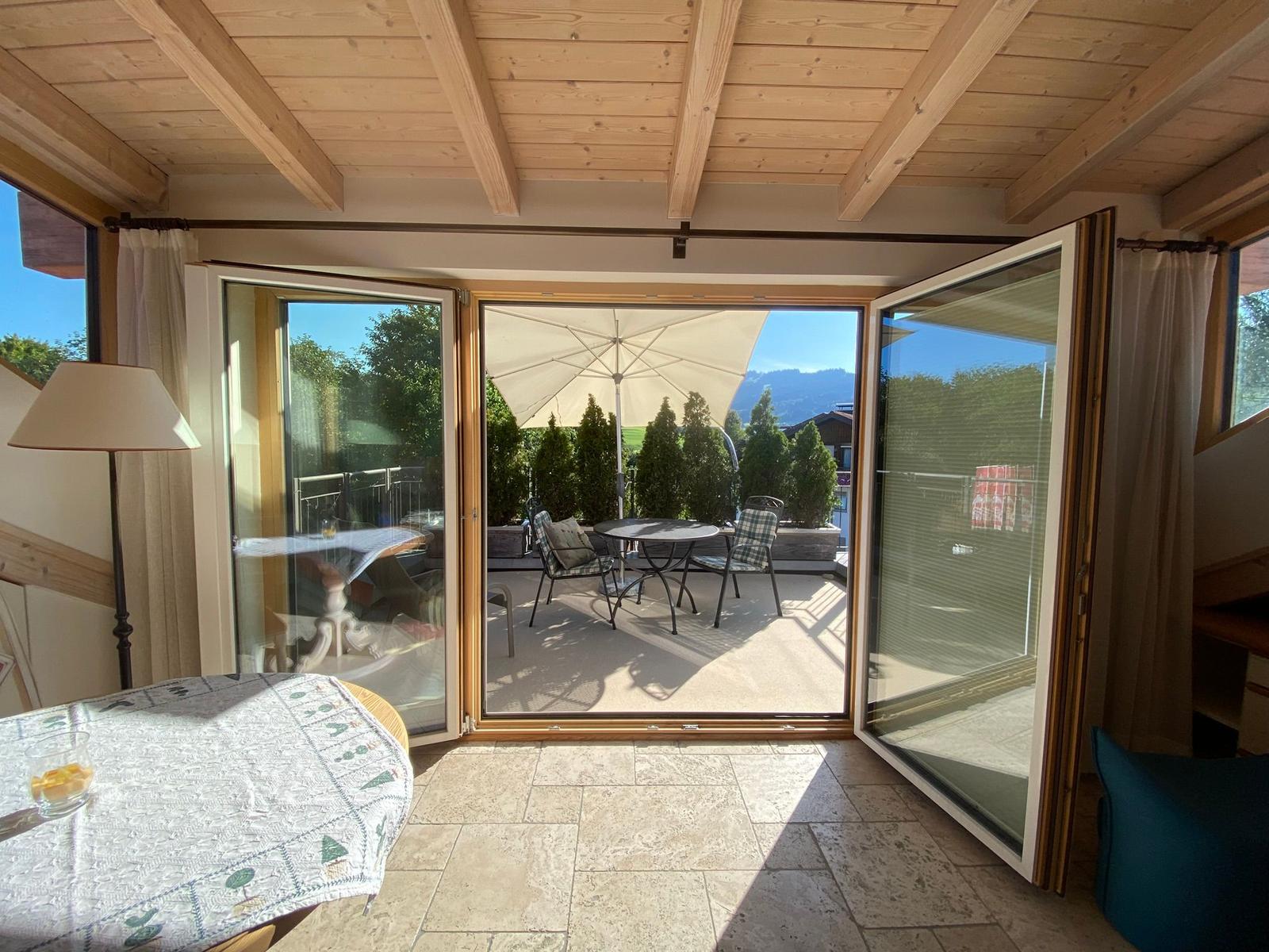 Terrasse mit Tisch, Stühlen und Sonnenschirm. Durchgehende Glasfront mit Holzrahmen.