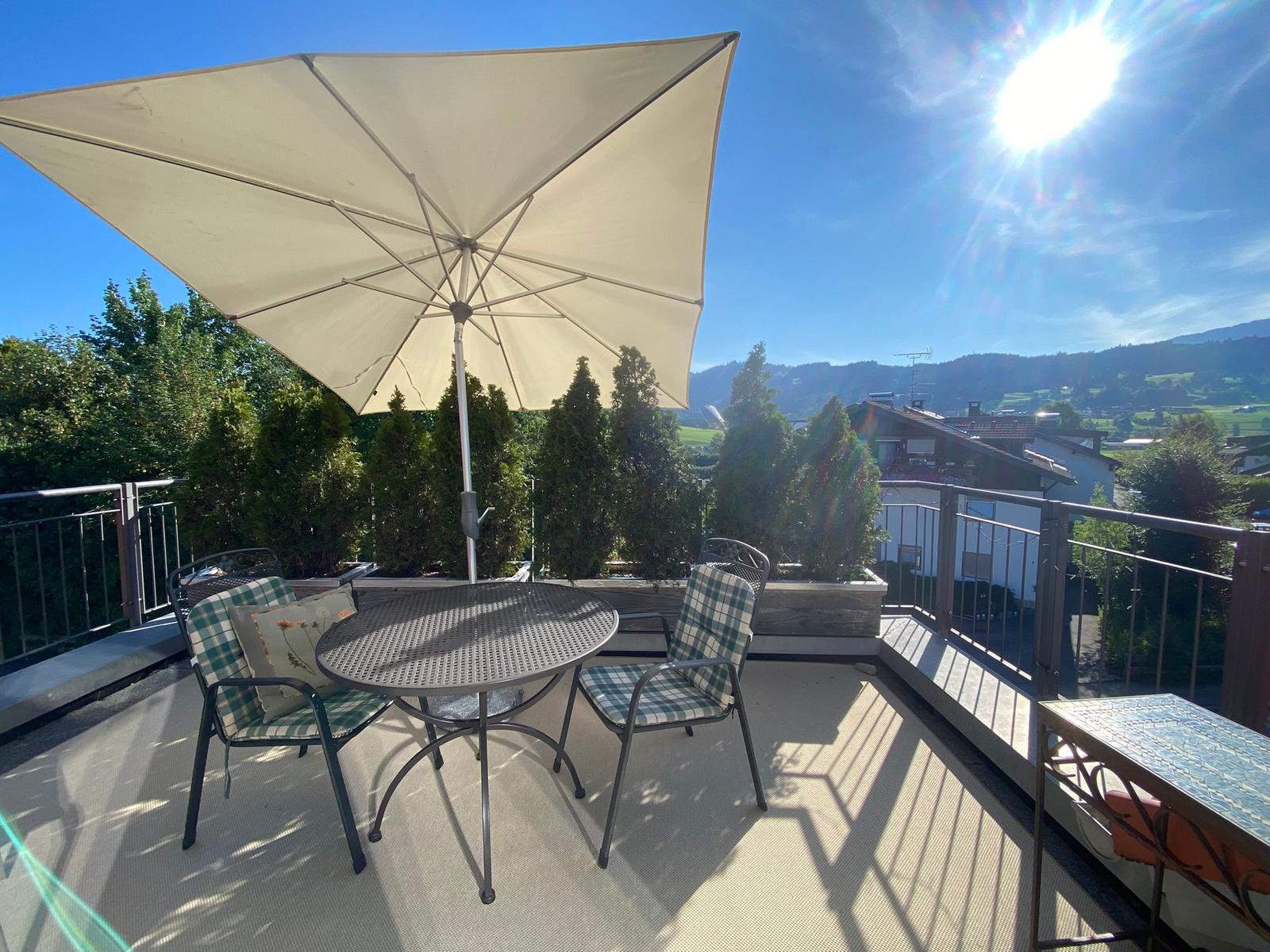 Terrasse mit Tisch, zwei Stühlen und Sonnenschirm. Blick auf grüne Hügel und Häuser.