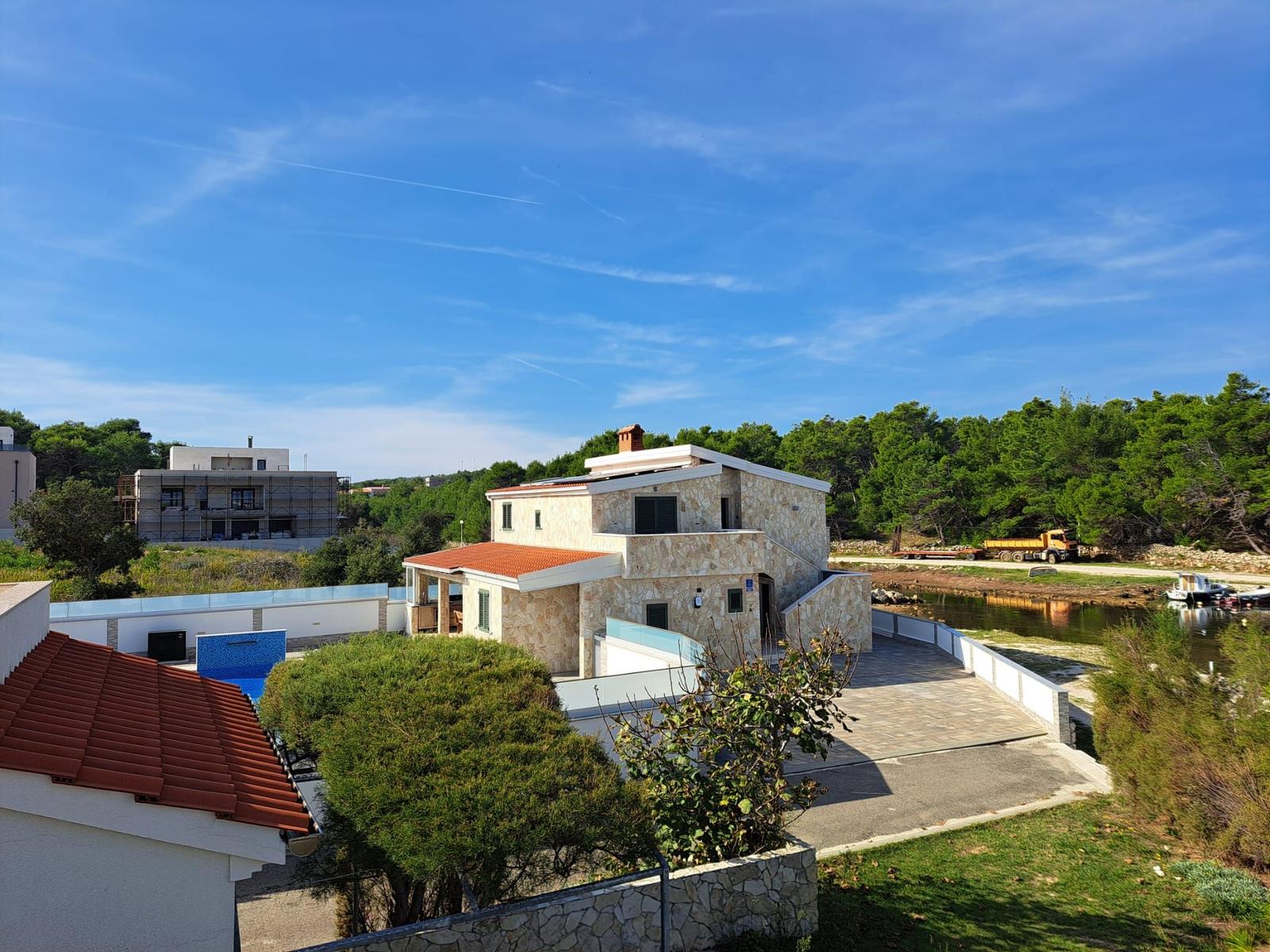 Villa mit Pool und Terrasse an Wasser, umgeben von Grün und klarer blauer Himmel.