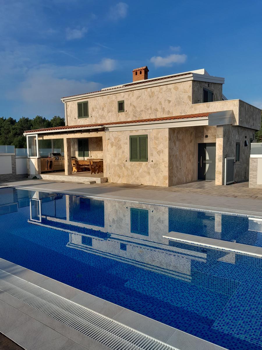 Villa mit Pool und Terrasse. Holztische und Stühle im Freien. Steinmauer und grüne Fensterläden.