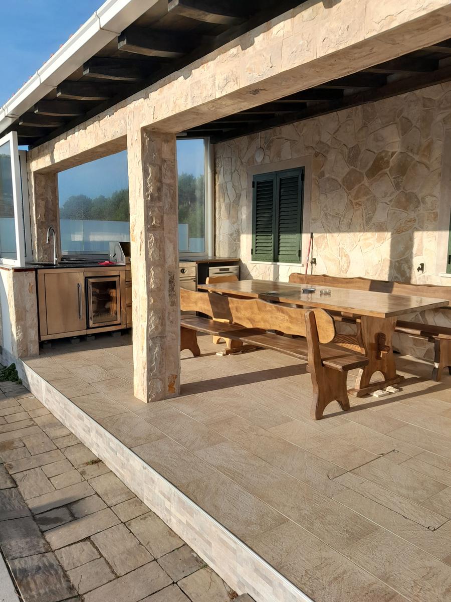 Terrasse mit Holztisch, Bänken und Grillbereich unter Steinmauer.