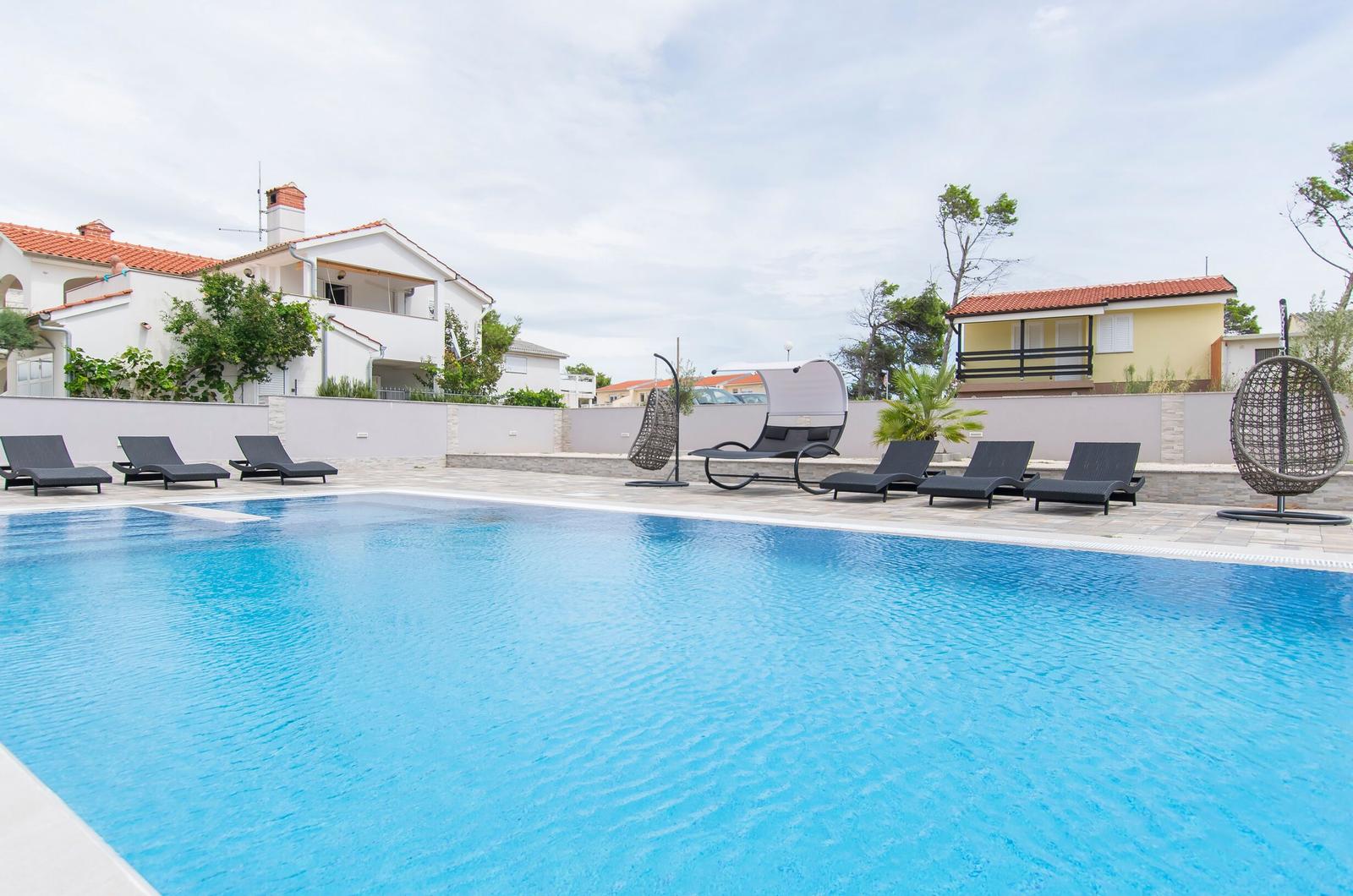 Pool mit Liegestühlen und Sonnenschirmen. Hintergrund: Wohnhäuser und Bäume.