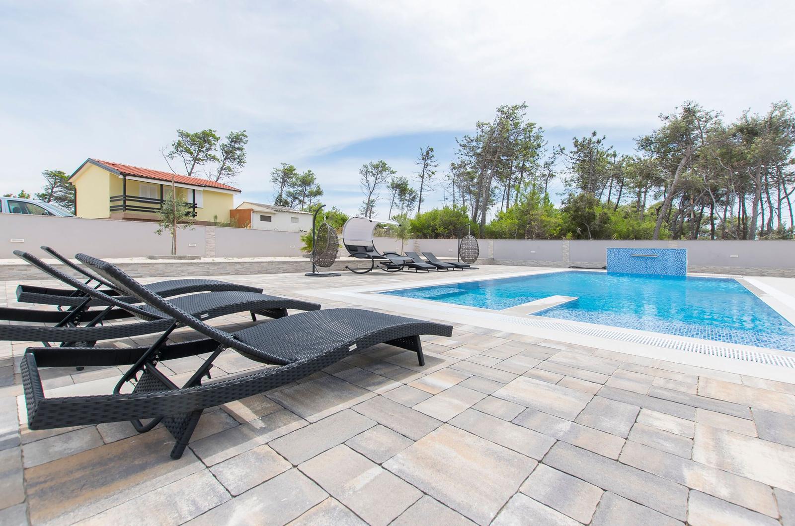 Poolbereich mit Liegestühlen und Garten. Hintergrund: Haus und Bäume.