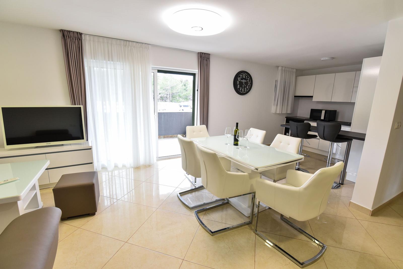 Esszimmer mit Tisch, Stühlen und Küchenbereich. Fernseher und Fenster mit Vorhängen.