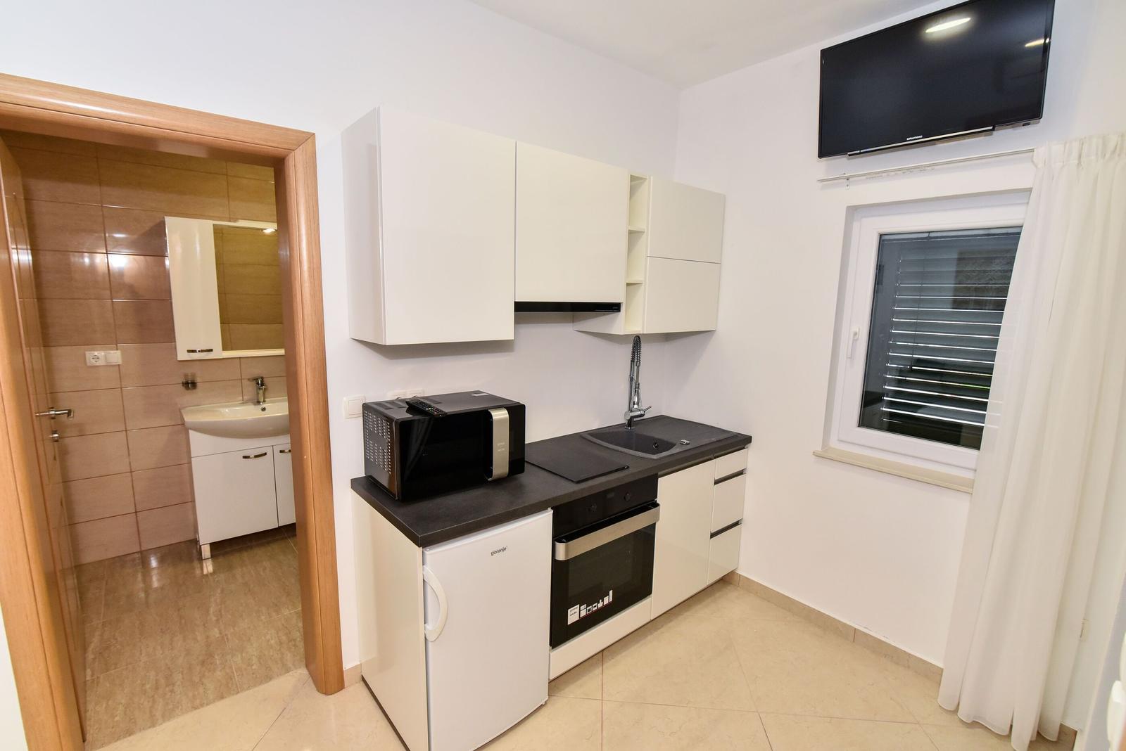 Küche mit Schränken, Spüle, Mikrowelle und Fenster. Im Hintergrund Badezimmer.