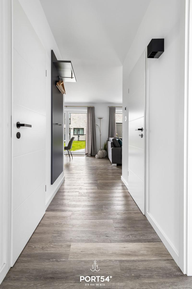 Flur mit grauem Holzboden, weißen Wänden und Tür zu Wohnzimmer mit Sofa und Gartenblick.
